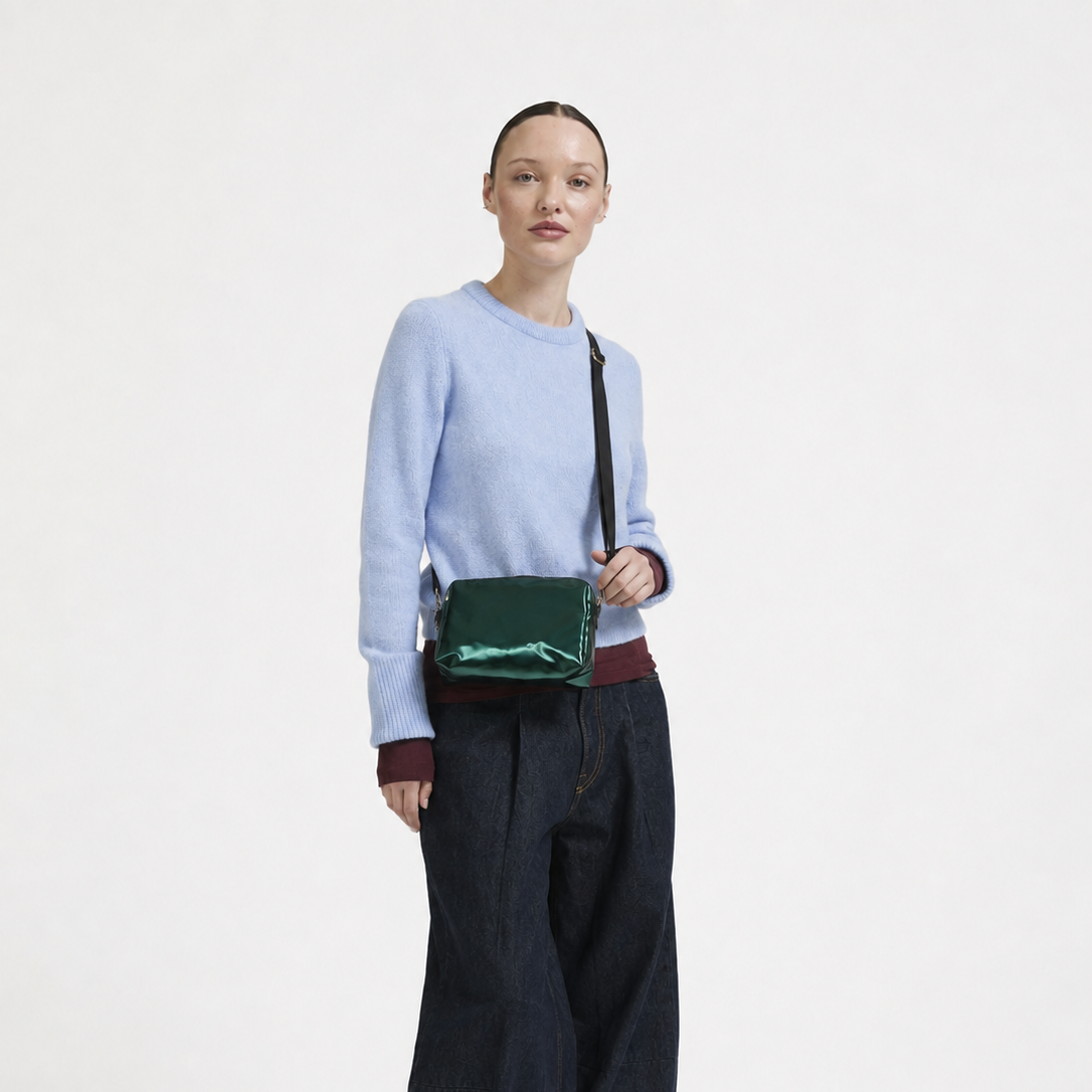 A model stands against a plain white studio background wearing a light blue knit sweater, dark wide-leg jeans, black shoes, and a shiny dark green crossbody bag.