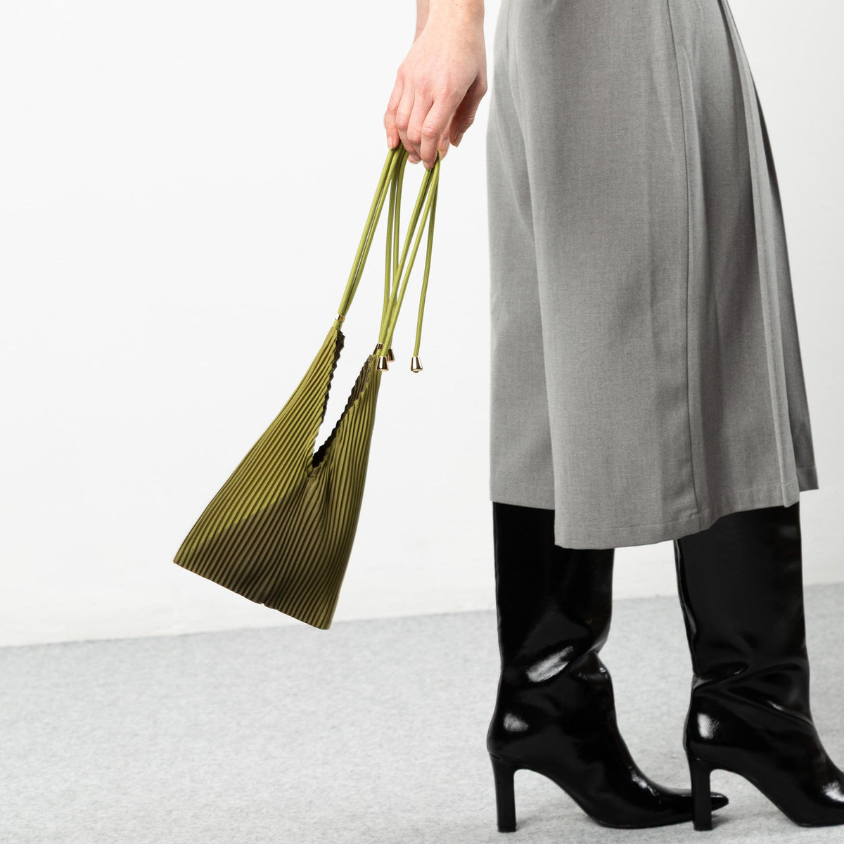 Person wearing a gray skirt and black shoes holding a green bag on a white background