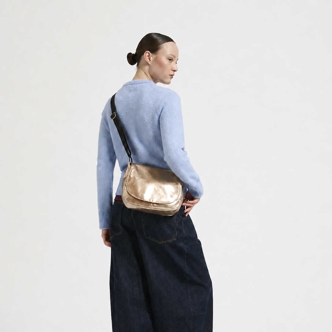 A model shown from the back wearing a light blue sweater, dark wide-leg denim jeans, and black shoes, while carrying a metallic gold crossbody bag with a black strap.