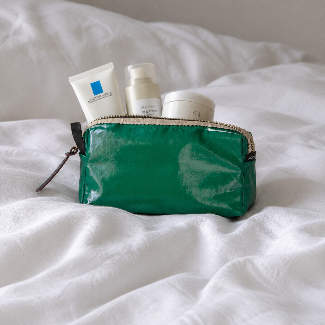 A green zippered cosmetic pouch filled with skincare items, placed on rumpled white bedding.