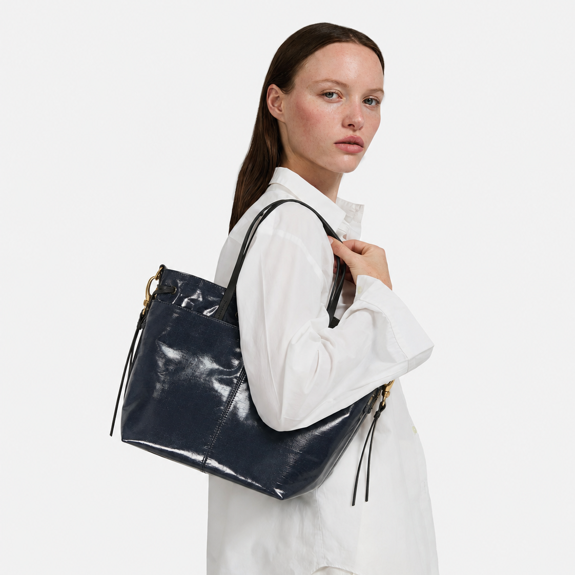 A woman wearing a white shirt and white pants carries a large glossy navy blue tote bag with gold-tone hardware and black drawstring tassels against a plain light gray studio background.