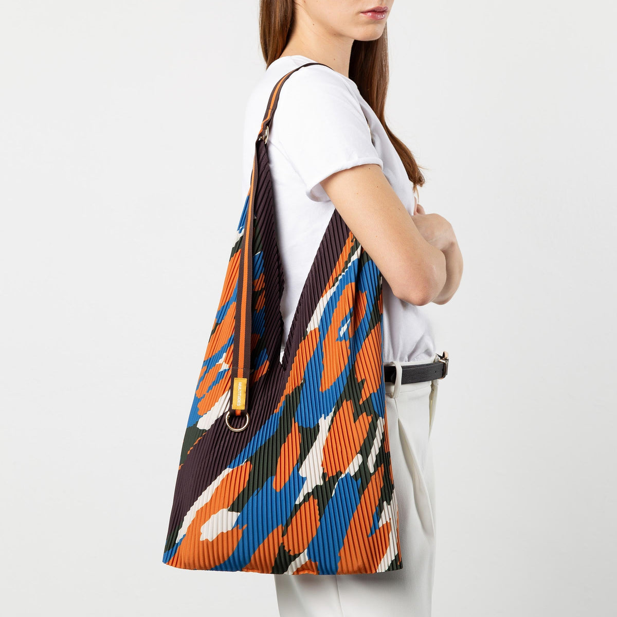 A woman wearing a white t-shirt and white trousers carries a large slouchy shoulder bag with a bold orange, blue, black, and white abstract pattern against a light gray studio background.