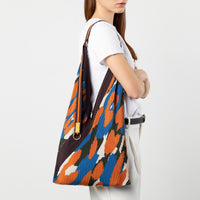 A woman wearing a white t-shirt and white trousers carries a large slouchy shoulder bag with a bold orange, blue, black, and white abstract pattern against a light gray studio background.